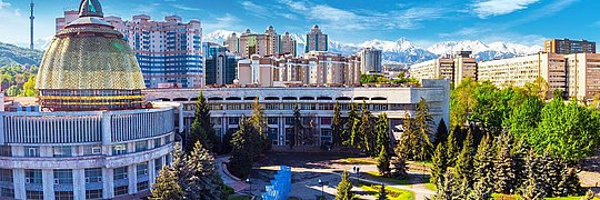 Städtische Landschaft mit modernen Gebäuden, darunter ein markantes Gebäude mit einer goldenen Kuppel. Im Hintergrund sind schneebedeckte Berge zu sehen, und im Vordergrund befinden sich grüne Parkanlagen mit Bäumen.