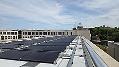 Dach mit mehreren Solarpaneelen auf einem flachen Gebäude vor bewölktem Himmel und Bäumen im Hintergrund