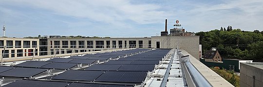 Dach mit mehreren Solarpaneelen auf einem flachen Gebäude vor bewölktem Himmel und Bäumen im Hintergrund