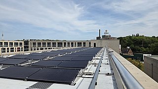 Dach mit mehreren Solarpaneelen auf einem flachen Gebäude vor bewölktem Himmel und Bäumen im Hintergrund
