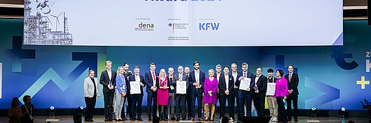 Gruppenfoto von Preisträgern des Energy Efficiency Award 2024 auf einer Bühne. Im Hintergrund steht ein großes Banner mit dem Text 'Energy Efficiency Award 2024' und Logos von dena und KFW. Die Personen halten Urkunden.