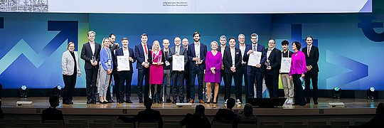 Gruppenfoto von Preisträgern des Energy Efficiency Award 2024 auf einer Bühne. Im Hintergrund steht ein großes Banner mit dem Text 'Energy Efficiency Award 2024' und Logos von dena und KFW. Die Personen halten Urkunden.