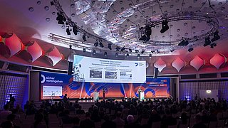 Großer Konferenzsaal mit Publikum, Bühne mit großem Bildschirm und roter, blauer Beleuchtung, Decke mit runden Leuchten und technischer Ausstattung.
