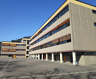 Mehrstöckiges Schulgebäude mit großen Fenstern und gelben Fensterläden unter klarem blauem Himmel