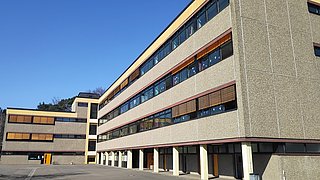 Mehrstöckiges Schulgebäude mit großen Fenstern und gelben Fensterläden unter klarem blauem Himmel