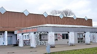 Blick auf ein flaches Gebäude mit Vordach am Eingang und braunem Flachdach, die Julius-Hirsch-Halle in Pfinztal