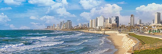 Küstenlinie von Tel Aviv mit Sandstrand und Wellen, im Hintergrund Hochhäuser unter blauem Himmel mit Wolken.