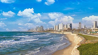 Küstenlinie von Tel Aviv mit Sandstrand und Wellen, im Hintergrund Hochhäuser unter blauem Himmel mit Wolken.