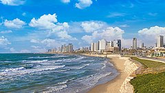 Küstenlinie von Tel Aviv mit Sandstrand und Wellen, im Hintergrund Hochhäuser unter blauem Himmel mit Wolken.