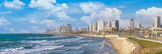 Küstenlinie von Tel Aviv mit Sandstrand und Wellen, im Hintergrund Hochhäuser unter blauem Himmel mit Wolken.