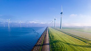 Landschaft mit Windkraftanlagen entlang eines Deichs, Wasser auf der linken Seite, grünes Feld auf der rechten Seite, blauer Himmel.