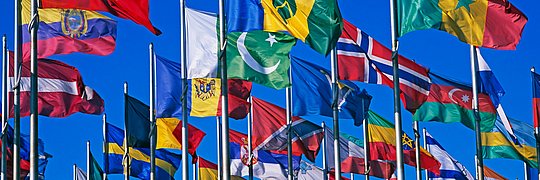 Mehrere Flaggen verschiedener Länder wehen an Fahnenmasten vor blauem Himmel