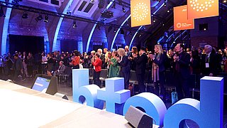 Menschenmenge applaudiert in einem Veranstaltungsraum mit blauen Buchstaben 'betd' im Vordergrund und orangefarbenen Bannern im Hintergrund.