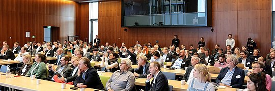 Eine große Gruppe von Menschen sitzt an Tischen in einem Konferenzraum und diskutiert miteinander.
