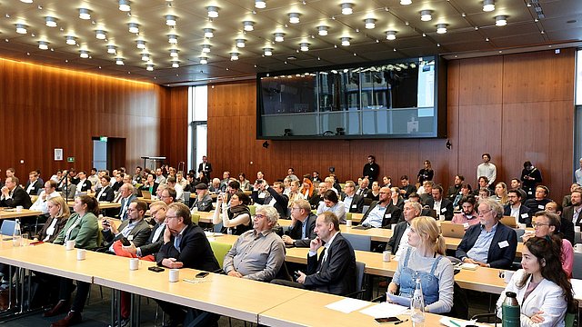 Eine große Gruppe von Menschen sitzt an Tischen in einem Konferenzraum und diskutiert miteinander.