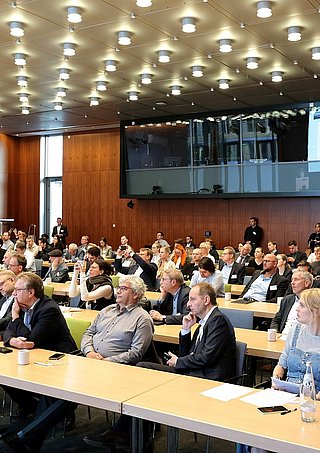 Eine große Gruppe von Menschen sitzt an Tischen in einem Konferenzraum und diskutiert miteinander.