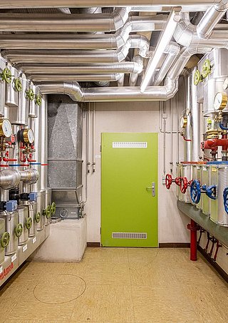 Blick in einen schmalen, hellen Kellerraum mit einer hellgrünen Tür am Raumende. Links und rechts sind Heizungsanlagen mit Wärmepumpen zu sehen