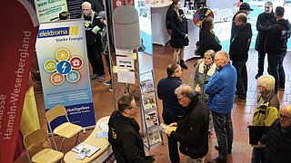Besucher der Woche der Wärmepumpe stehen an verschiedenen Ständen, darunter ein Stand mit einem Plakat der Firma „elektroma“ und Informationsmaterialien.