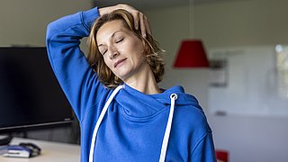 Eine Person mit blonden Haaren trägt einen blauen Hoodie und hat eine Hand hinter dem Kopf. Im Hintergrund sind ein Computerbildschirm und eine rote Lampe sichtbar.