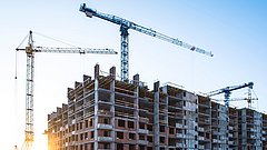 Der Sonnenuntergang wirft Licht auf eine Baustelle mit Kränen und einem teilweise fertiggestellten mehrstöckigen Gebäude mit Beton- und Ziegelstrukturen.