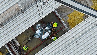 Drohnenfoto, Blick ins offene Dach mit Handwerkern und Kran der Rohre in das Dach transportiert.