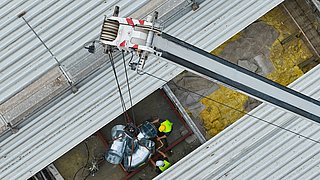 Drohnenfoto, Blick ins offene Dach mit Handwerkern und Kran der Rohre in das Dach transportiert.