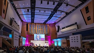 Großer Konferenzsaal mit Publikum und Podium vor einer großen Orgel und einer Leinwand mit einer Frau, die spricht