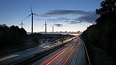 Dämmerungsaufnahme einer Autobahn mit fließendem Verkehr. Im Hintergrund stehen mehrere Windkraftanlagen und der Himmel ist bewölkt.