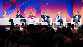 Fünf Personen sitzen auf weißen Sesseln auf einer Bühne vor einem Publikum, bunte Wand mit Pfeilmustern im Hintergrund.