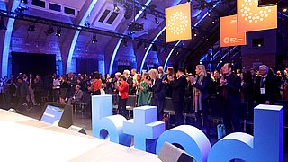 Menschenmenge applaudiert in einem Veranstaltungsraum mit blauen Buchstaben 'betd' im Vordergrund und orangefarbenen Bannern im Hintergrund.