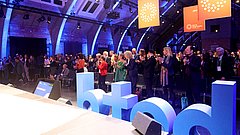 Menschenmenge applaudiert in einem Veranstaltungsraum mit blauen Buchstaben 'betd' im Vordergrund und orangefarbenen Bannern im Hintergrund.