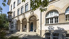 Eingang der Theodor-Heuss-Realschule mit drei Säulen und einem Treppenaufgang. Fenster mit weißen Rahmen und blauen Türen.