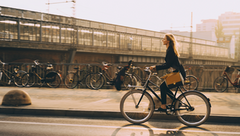 Frau fährt Fahrrad auf einer Straße neben einem Fahrradparkplatz mit mehreren abgestellten Fahrrädern im Hintergrund.
