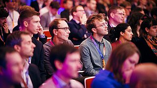 Mehrere Personen sitzen in einem Publikum, einige tragen Ausweishalter um den Hals, die Kleidung ist überwiegend formell bis casual.