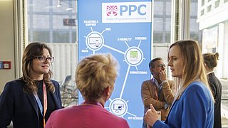 Vier Personen im Gespräch vor einem Display mit dem Logo 'PPC Power Plus Communications' und einer schematischen Darstellung von E-Mobility-Komponenten in einem hellen, modernen Innenraum.