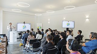 Simon Kreye, Ständiger Vertreter und Leiter des Wirtschaftsreferats der deutschen Botschaft in Vietnam, hält einen Vortrag vor Publikum in einem modernen Besprechungsraum. Im Raum stehen zwei moderne Bildschirme und ein Roll-up, dort ist jeweils eine stilisierte Weltkarte und die Schrift "Climate Talks Hanoi" zu sehen.