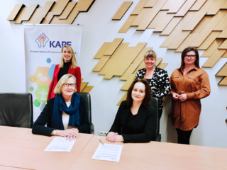 Fünf Personen in einem Büro bei der Unterzeichnung einer Kooperationsvereinbarung zwischen KAPE und dena, zwei sitzen am Tisch mit Dokumenten, drei stehen dahinter vor einer KAPE-Werbetafel.