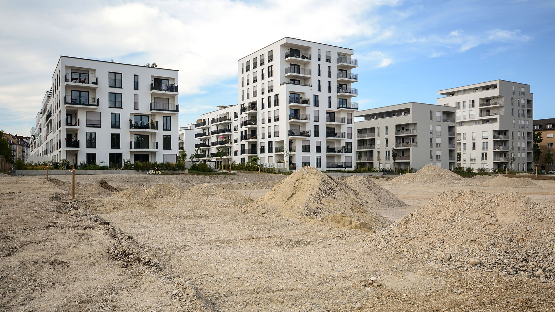 Moderne Wohngebäude mit mehreren Etagen, Balkonen und großen Fenstern, umgeben von unbebautem Gelände mit Erdhügeln.