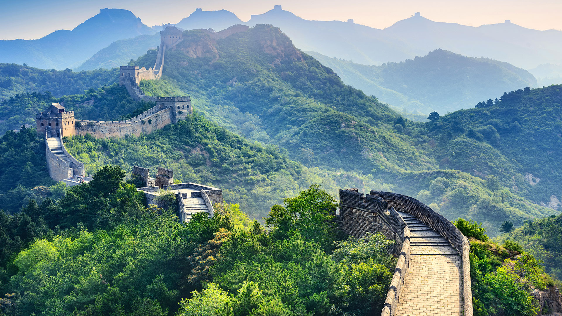 Die Chinesische Mauer erstreckt sich über grüne Hügel, umgeben von Bäumen und Bergen im Hintergrund. Die Mauer ist aus Stein und Ziegeln gebaut.