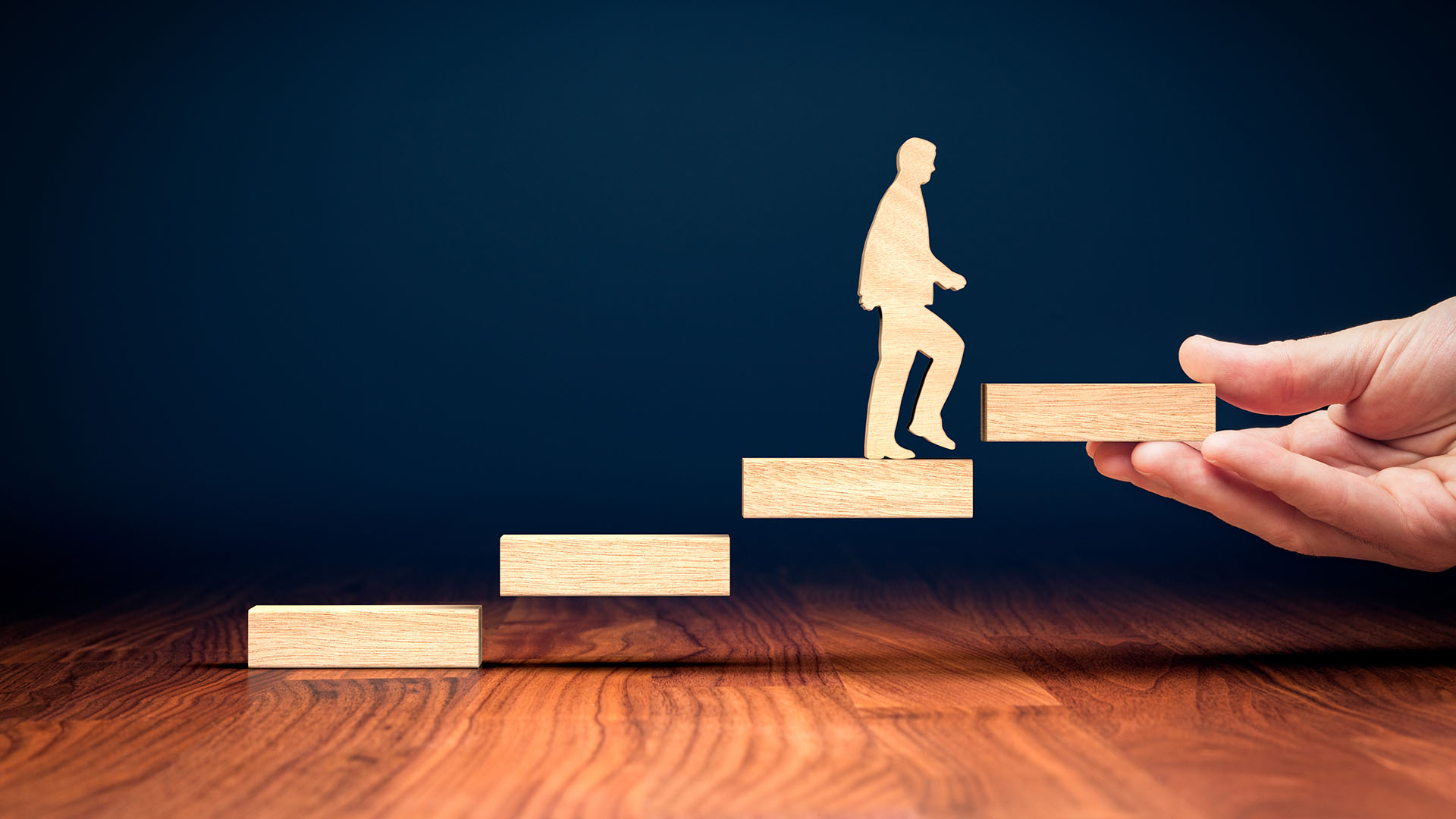 Eine Hand hält eine Treppe aus Holzblöcken, auf der eine stilisierte Figur nach oben steigt.