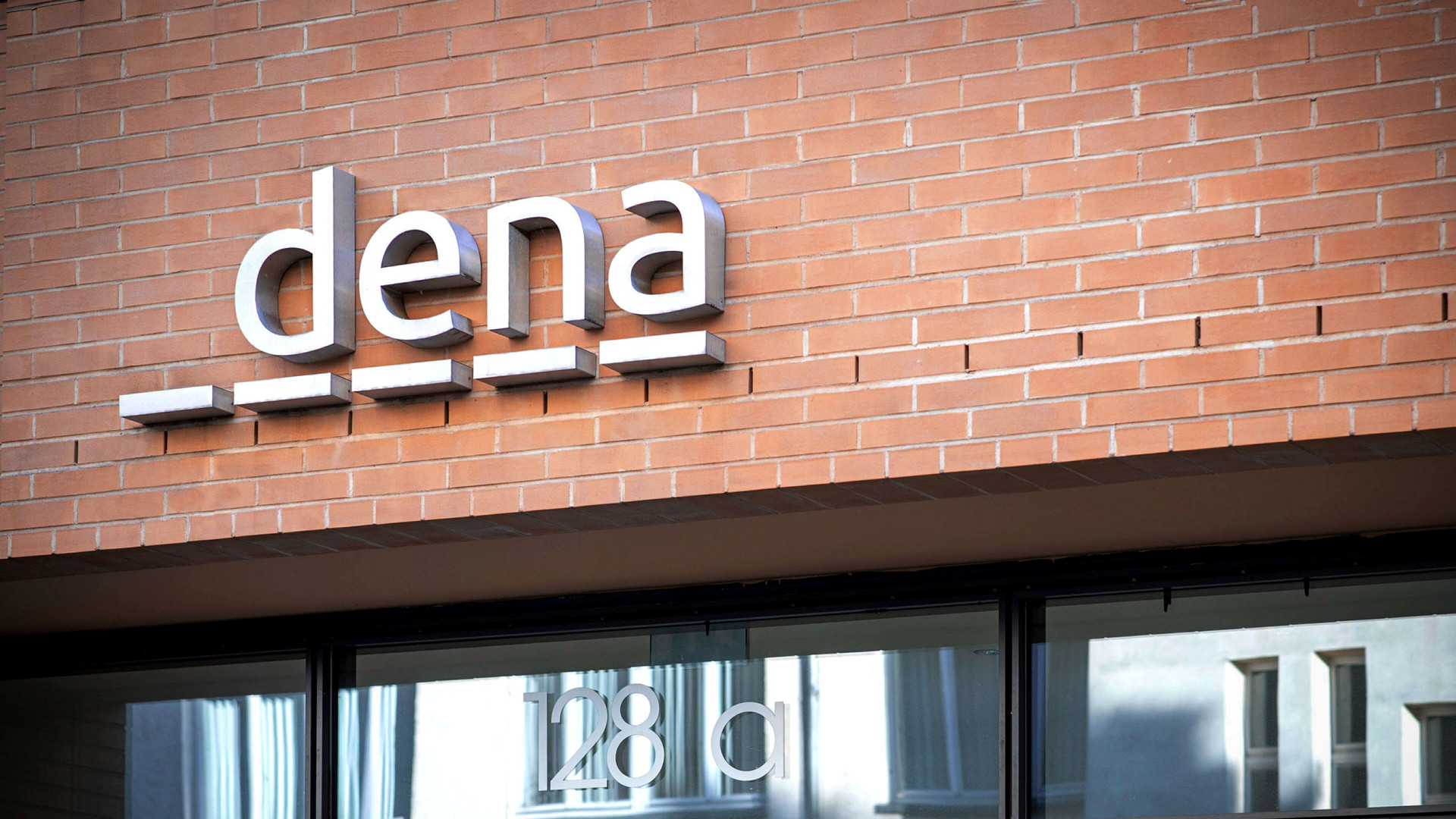 Logo von dena in moderner Schriftart, montiert an einer Ziegelwand. Darunter steht die Hausnummer '128 a'.