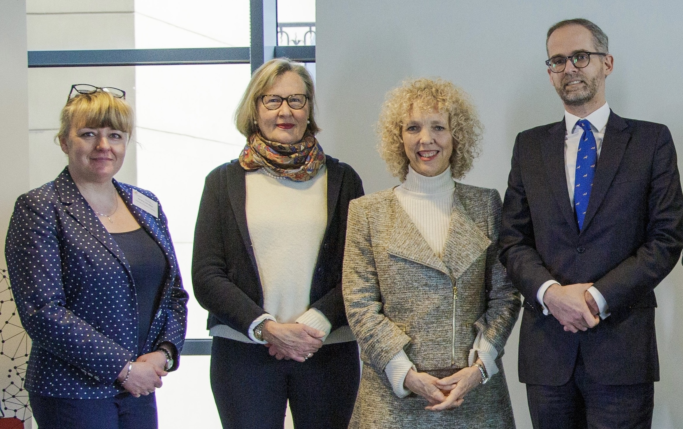 Vier Personen stehen nebeneinander in einem hellen Raum, drei Frauen und ein Mann in formeller Kleidung, Hände vor dem Körper gefaltet