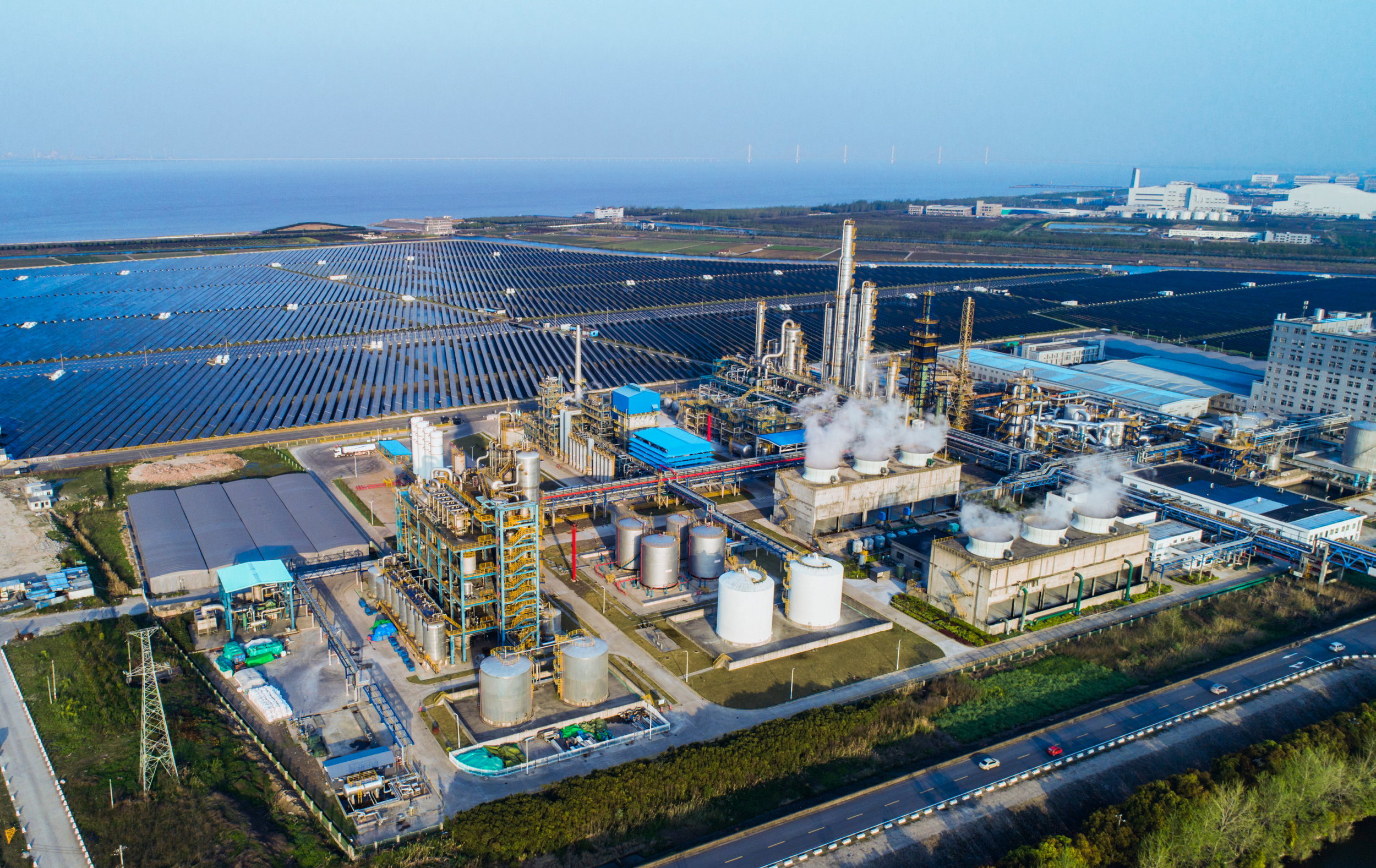 Industrieanlage mit rauchenden Kühltürmen, umgeben von großen Solarpanel-Feldern nahe der Küste.