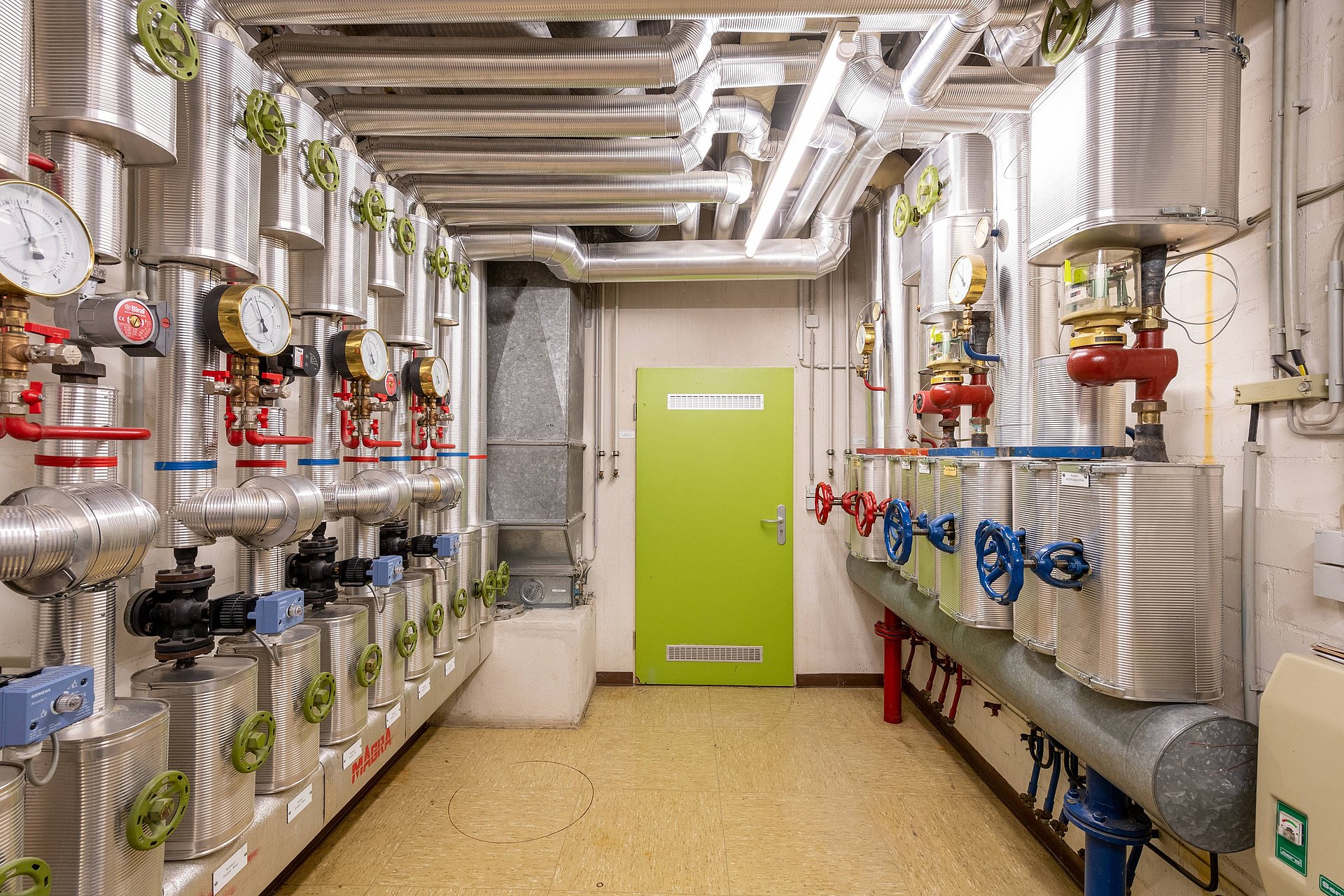 Blick in einen schmalen, hellen Kellerraum mit einer hellgrünen Tür am Raumende. Links und rechts sind Heizungsanlagen mit Wärmepumpen zu sehen