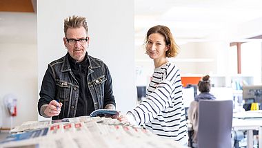 Zwei Personen stehen in einem Büro mit Zeitungen.