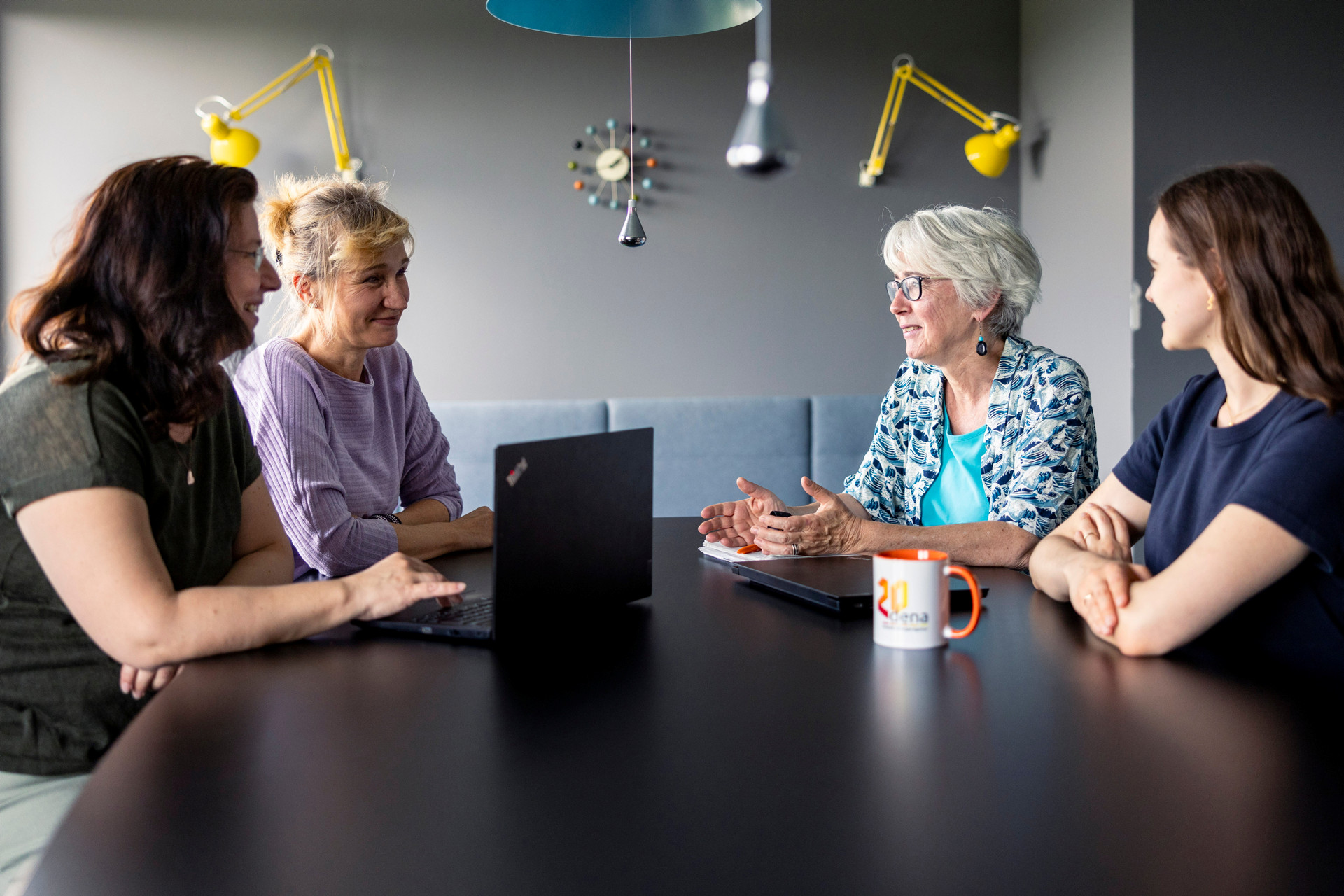 Vier Personen sitzen an einem Konferenztisch, zwei haben Laptops vor sich, eine hält eine Tasse mit dem 'dena' Logo, im Hintergrund zwei gelbe Lampen an der Wand
