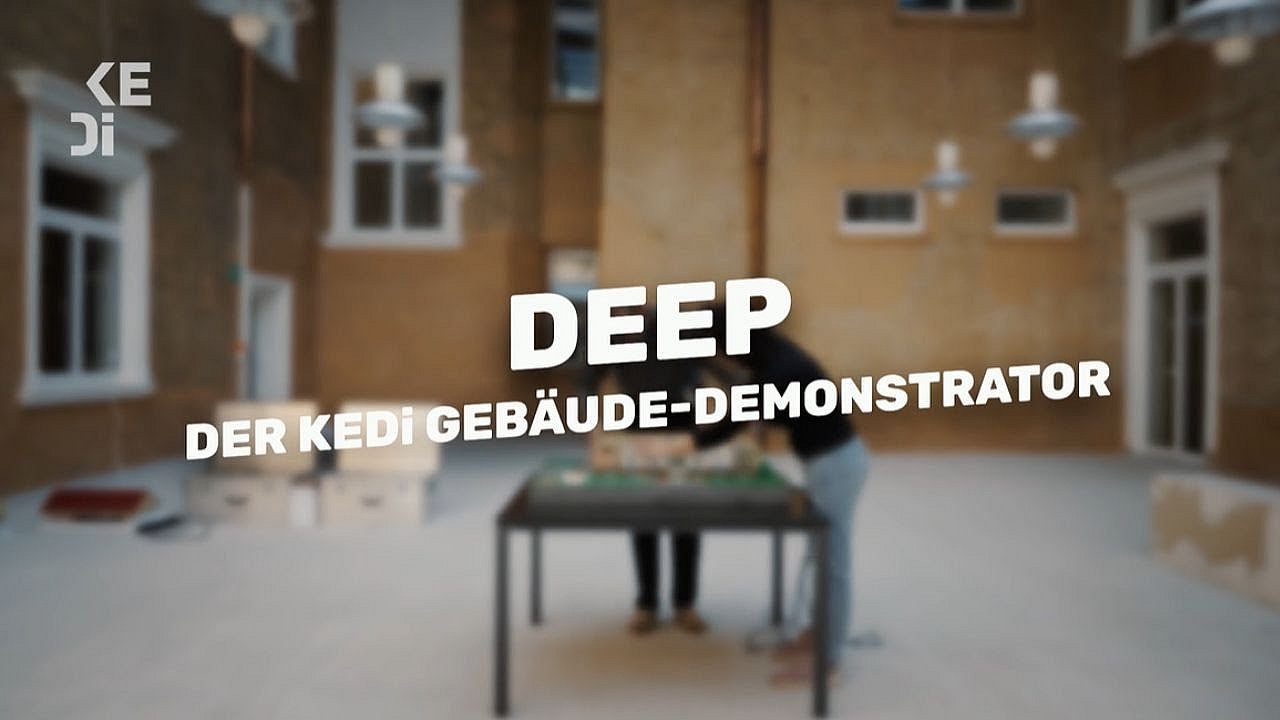 Zwei Personen arbeiten an einem Tisch in einem offenen Raum mit Backsteinwänden. Text: 'DEEP – Der KEDi Gebäude-Demonstrator'.